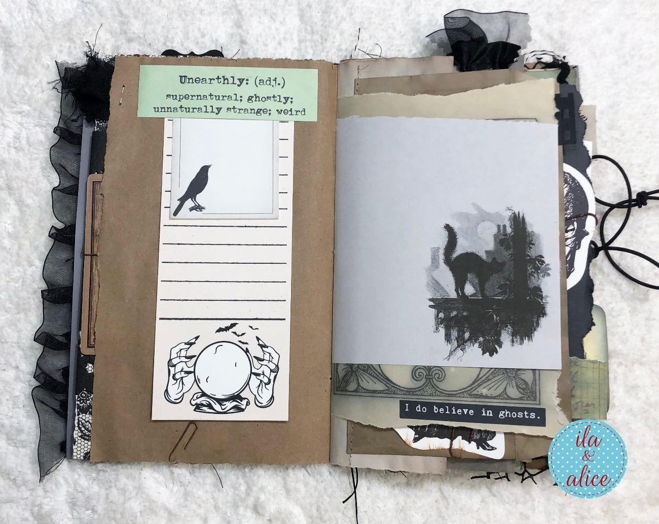 An open handmade scrapbook journal with a gothic collage layout, featuring black ruffled trim, layered paper layers, printed text, silhouette artwork of a cat and crow, and a small sticker reading 'I do believe in ghosts.' photographed on a white textured fabric background.