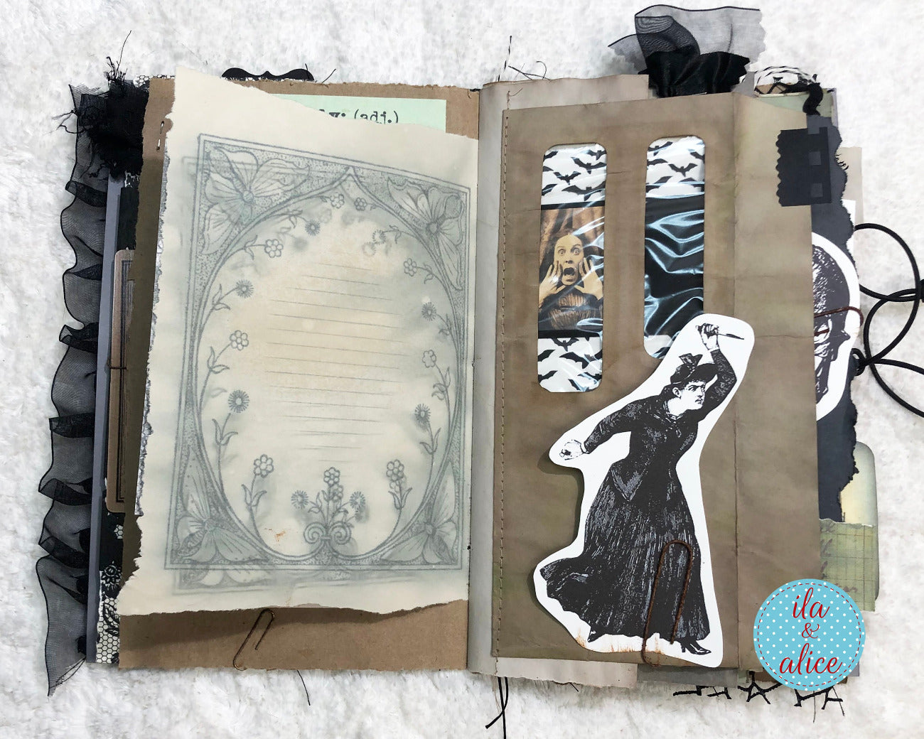 An open handmade scrapbook journal or art book on a white textured fabric background. The pages feature vintage-style elements including a floral frame, torn paper, black ruffled trim, patterned washi tape, a die-cut black dress figure, and small photo inserts.
