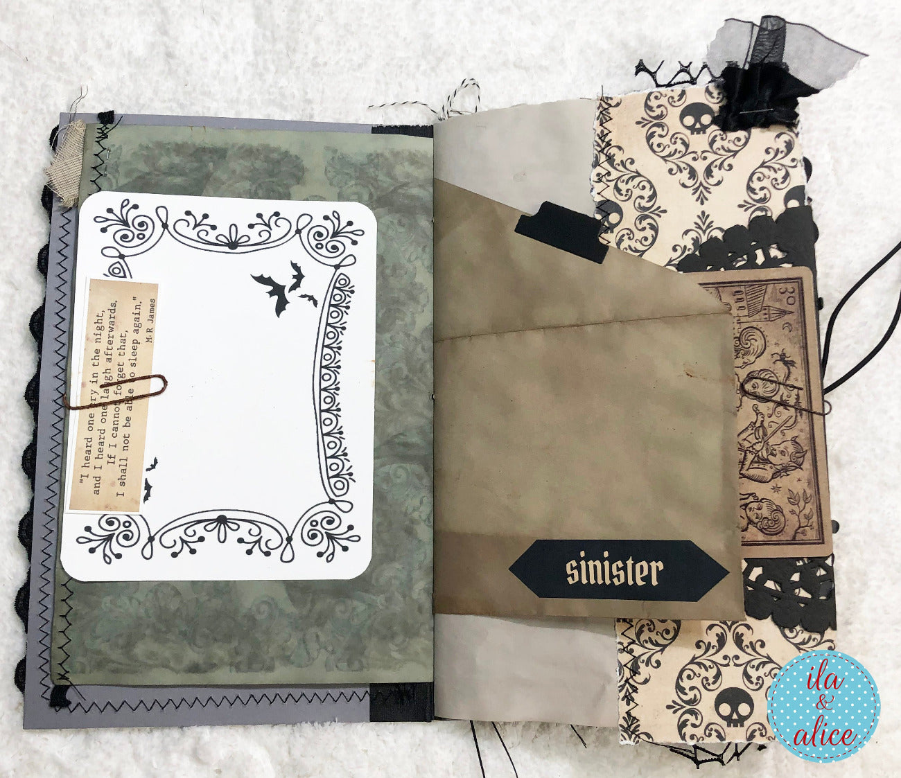 An open handmade scrapbook journal or art book with layered vintage-style pages, black lace, paper clips, and gothic illustrations. A white insert card with an ornate black frame and small bat silhouettes is attached on the left, and tan paper tabs, black labels, and patterned paper are layered on the right.