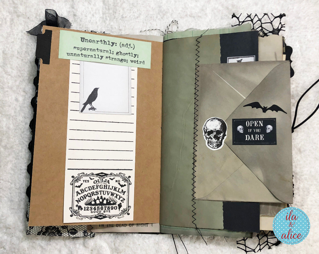 An open handmade scrapbook journal with gothic-themed pages, featuring a bird illustration, a Ouija board design, bat silhouettes, a skull sticker, and a lace bookmark on a white textured background.