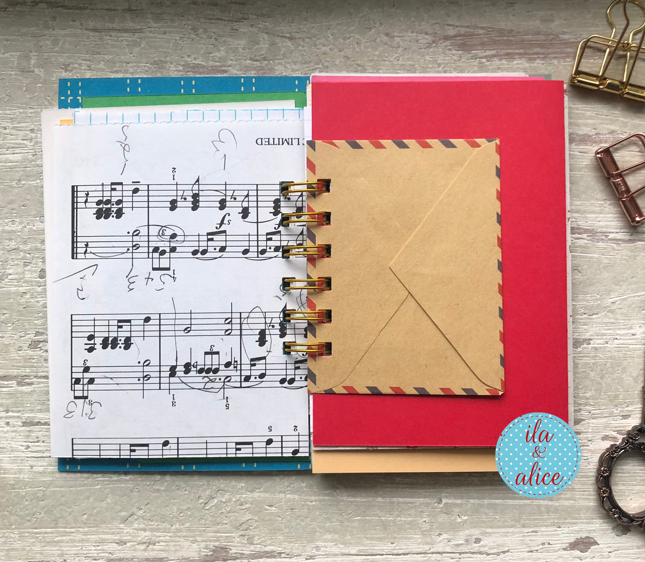 Red notebook with a brown envelope on top, music sheet on the left, on a wooden surface.