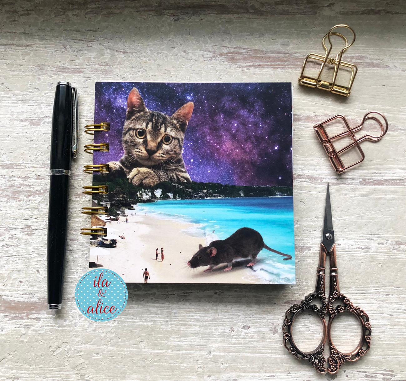 Notebook with cat and mouse illustration on a beach, surrounded by stationery items on a textured surface.