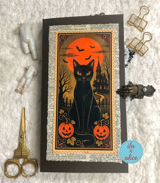 A Halloween-themed art print or card featuring a black cat in front of an orange full moon, displayed on a textured white fabric background with decorative scissors, binder clips, and small figurines.