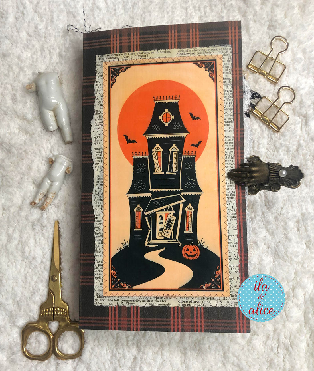 A Halloween-themed card or art print featuring a black haunted house silhouette, an orange full moon, bats, and a jack-o'-lantern, styled on a white textured fabric with small decorative props including scissors, binder clips, figurines, and a claw clip.