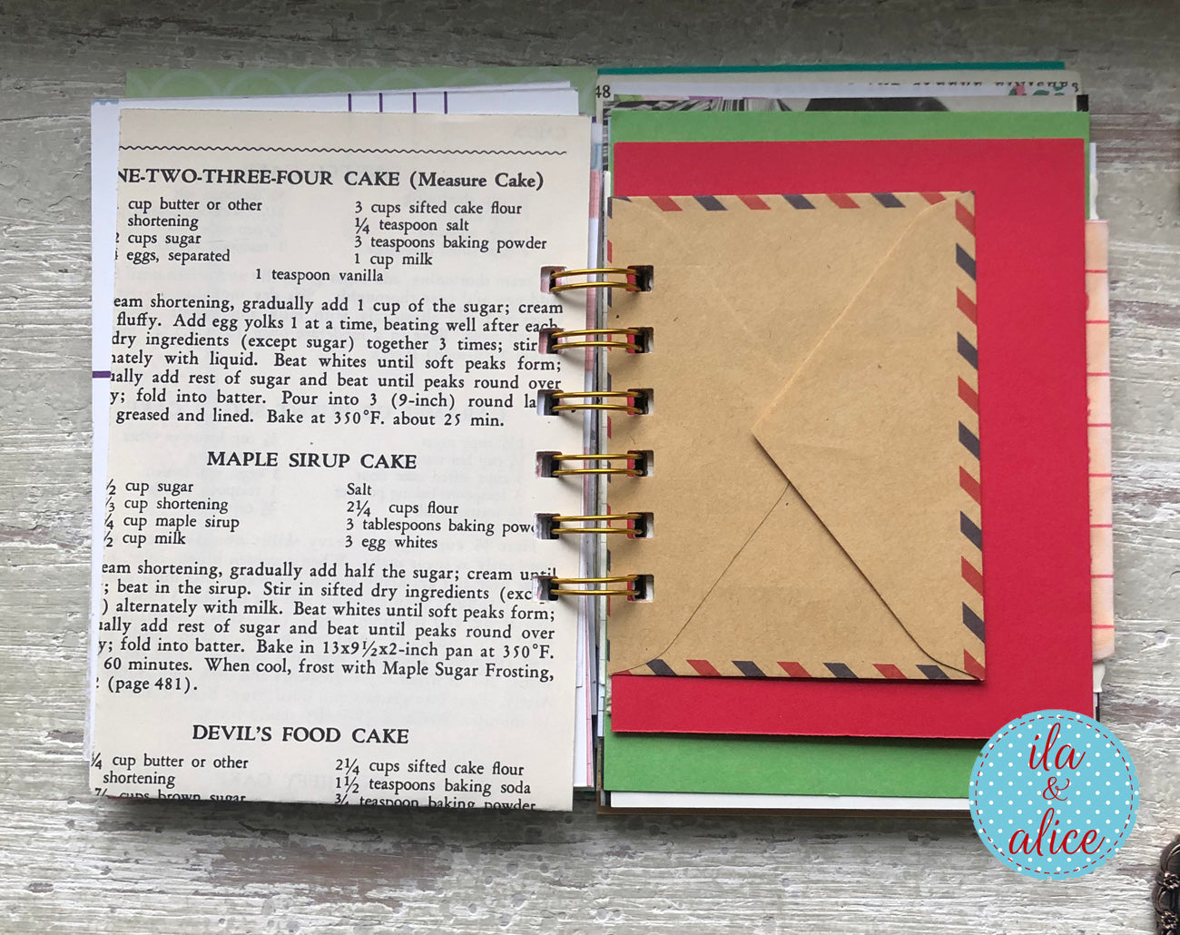 Notebook with recipe pages and a letter on a wooden surface, featuring the brand 'ila & alice'.