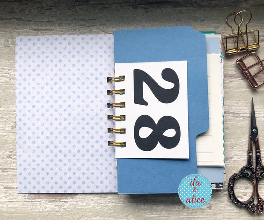 Blue notebook with a sticker displaying the number 28, surrounded by office supplies on a wooden surface.