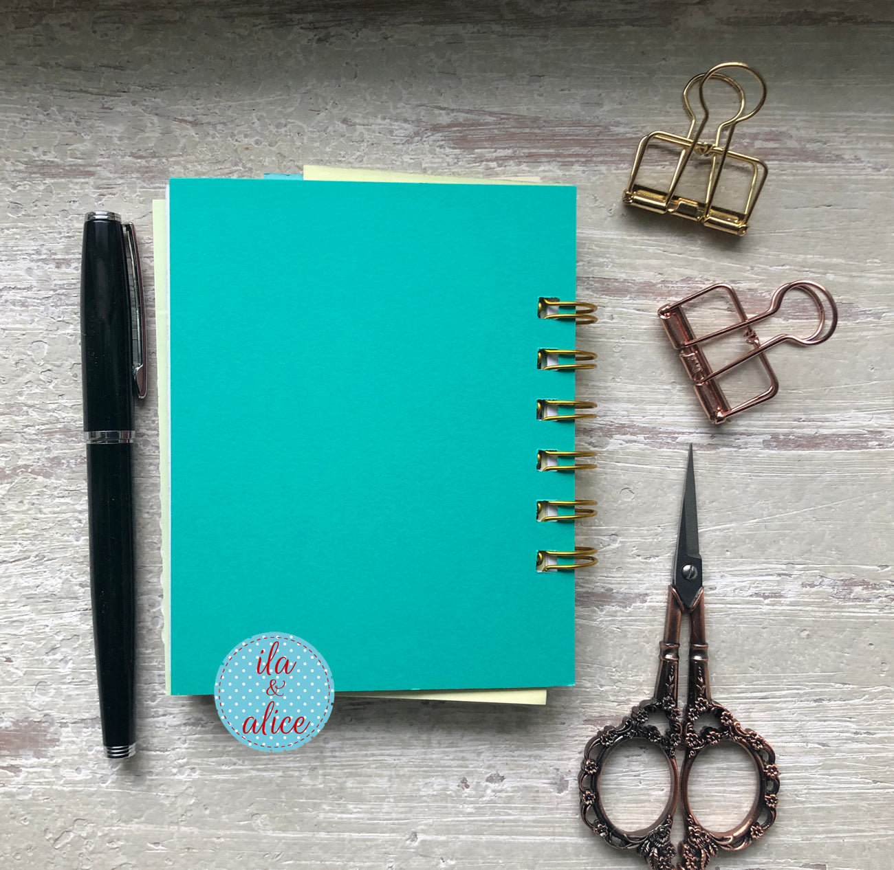 Turquoise notebook with a pen, scissors, and paper clips on a gray surface.