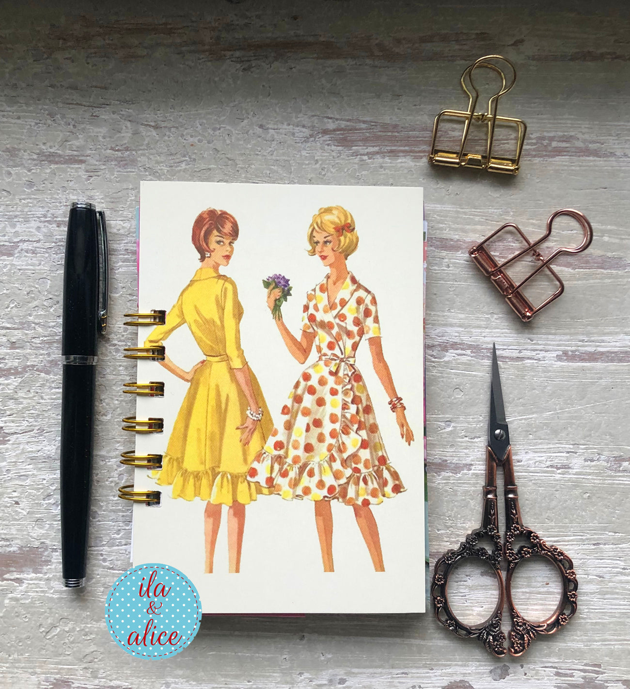 Notebook with vintage-style illustrations of two women, pen, scissors, and paper clips on a wooden surface.