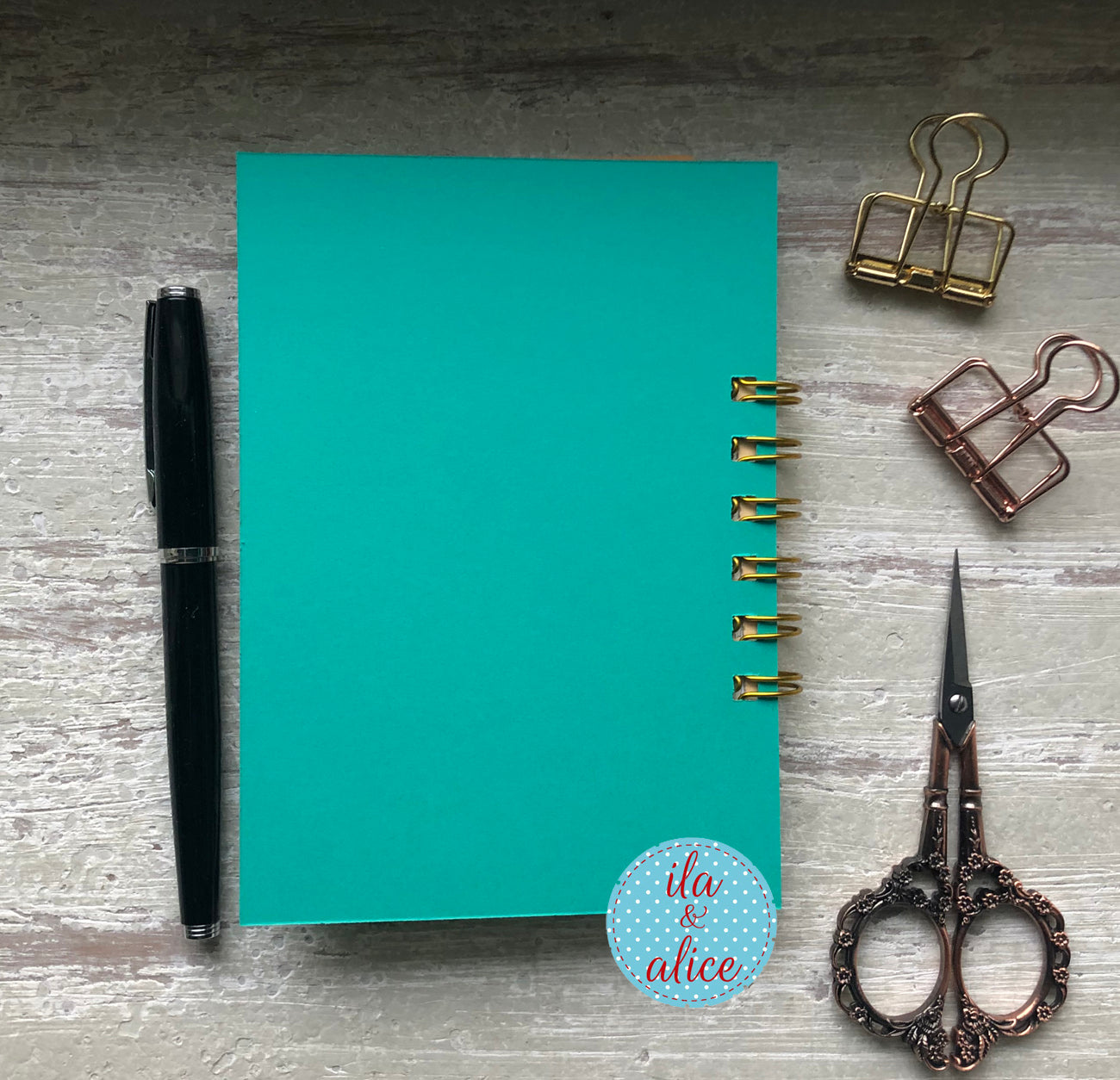 Turquoise notebook with stationery items on a wooden surface, featuring the brand 'ila & alice'.